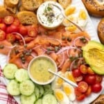 a smoked salmon charcuterie style platter with cucumbers, tomatoes, hard boiled eggs, dill mustard sauce, and cream cheese. Next to the platter are everything bagels.