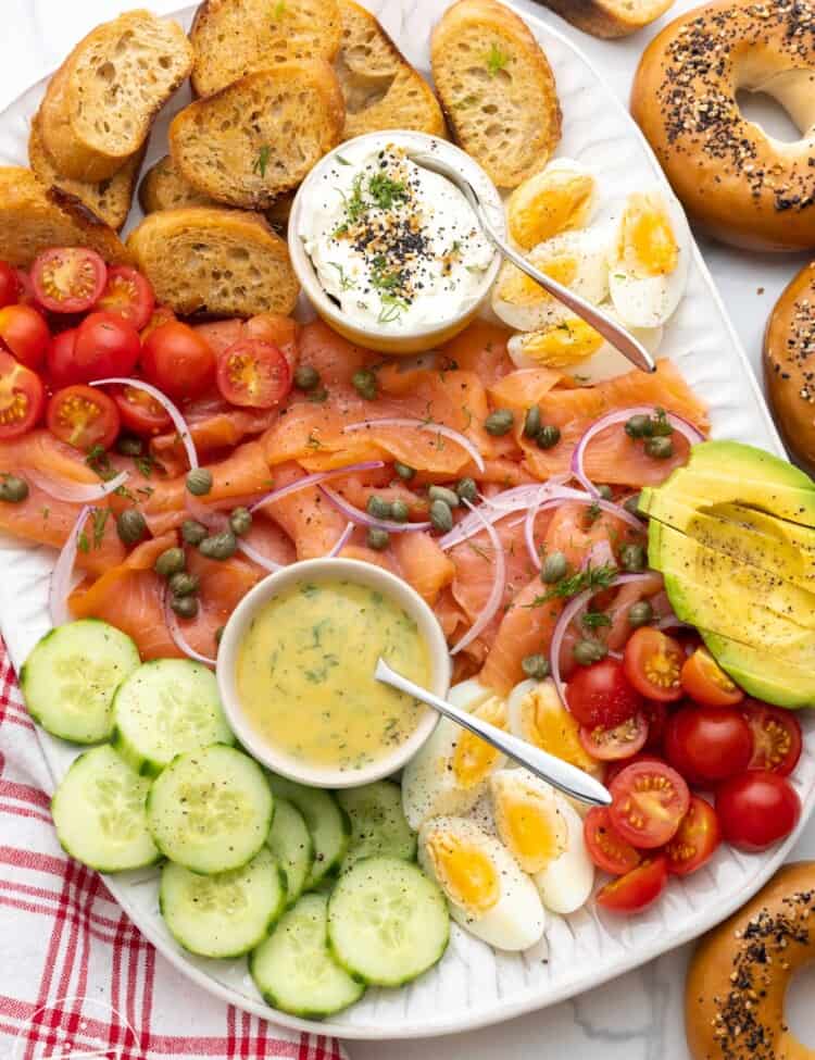 a smoked salmon charcuterie style platter with cucumbers, tomatoes, hard boiled eggs, dill mustard sauce, and cream cheese. Next to the platter are everything bagels.