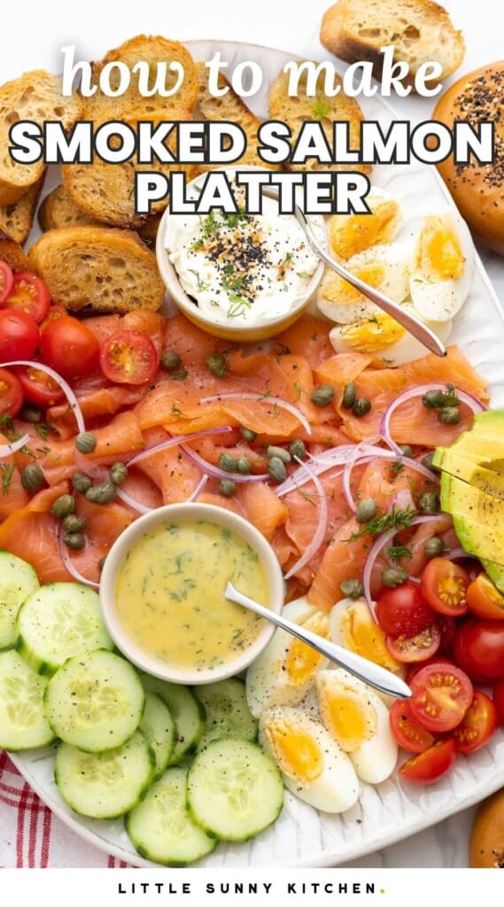 a white platter of smoked salmon topped with onions and capers. Around the salmon are small bowls of sauces, sliced tomatoes and cucumbers, hard boiled eggs, bagels, and homemade crostini.