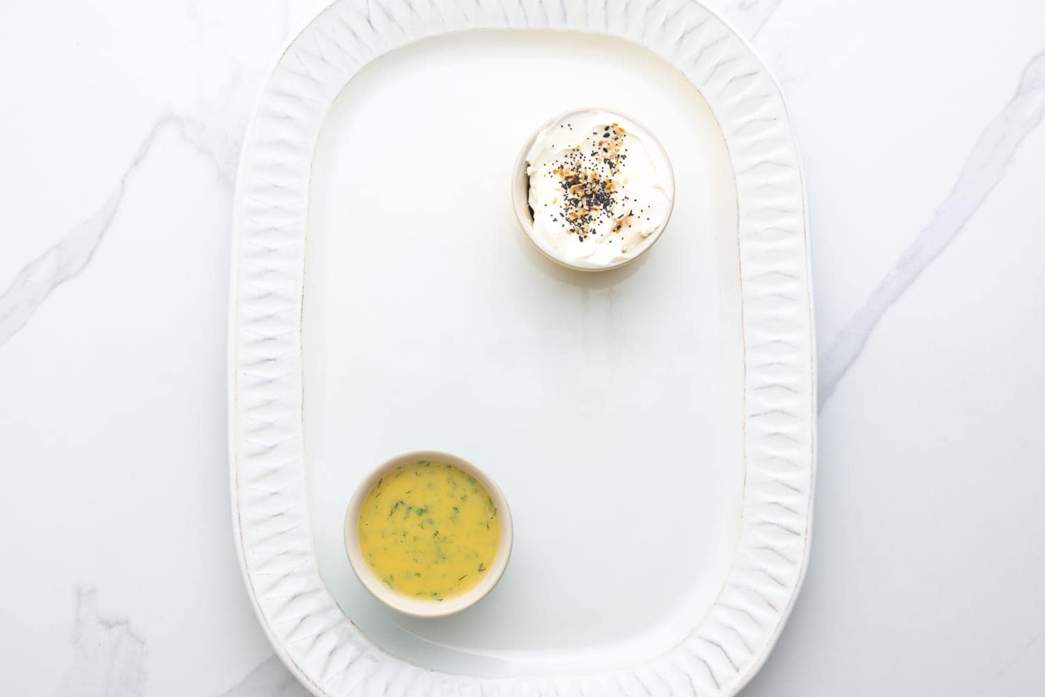 A rounded rectangular platter holding a small bowl of cream cheese with everything bagel seasoning and a small bowl of dill mustard sauce.