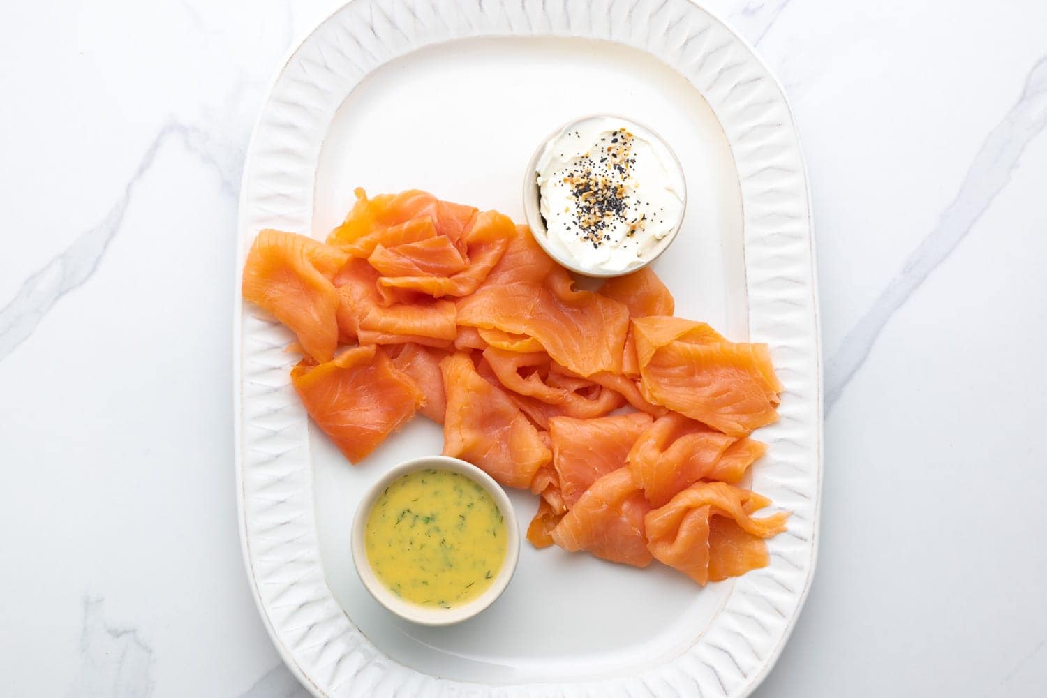 Slices of smoked salmon added to a white ceramic platter with sauces.