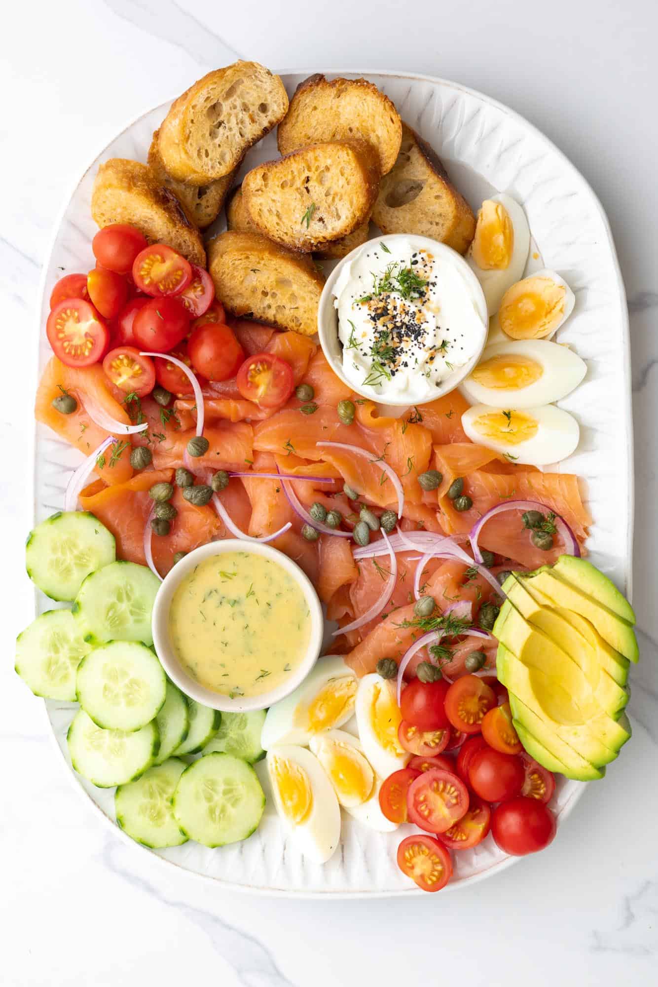 a filled rectangular white platter holding artfully arranged smoked salmon and toppings for bagels or crostini.