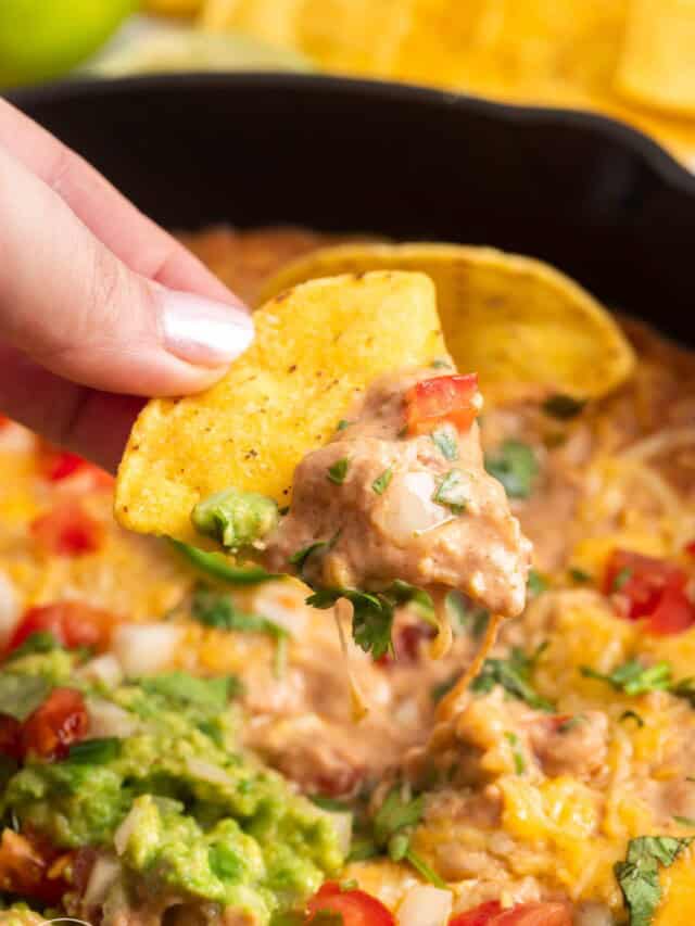 a hand holding a tortilla chip that has been dipped into cream cheese bean dip with pico de gallo and guac.