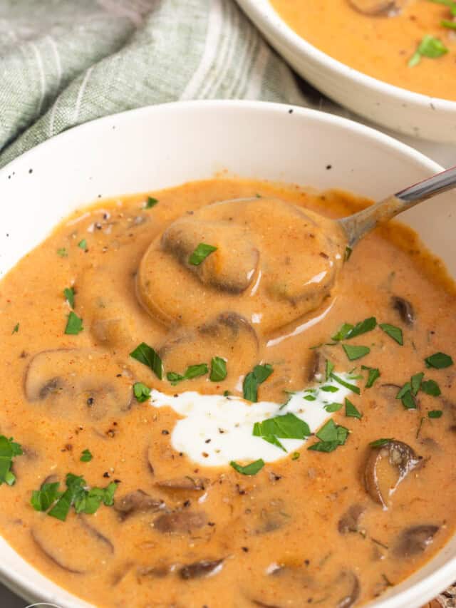 a white bowl of brown creamy soup with mushrooms. A spoon is taking a bite.