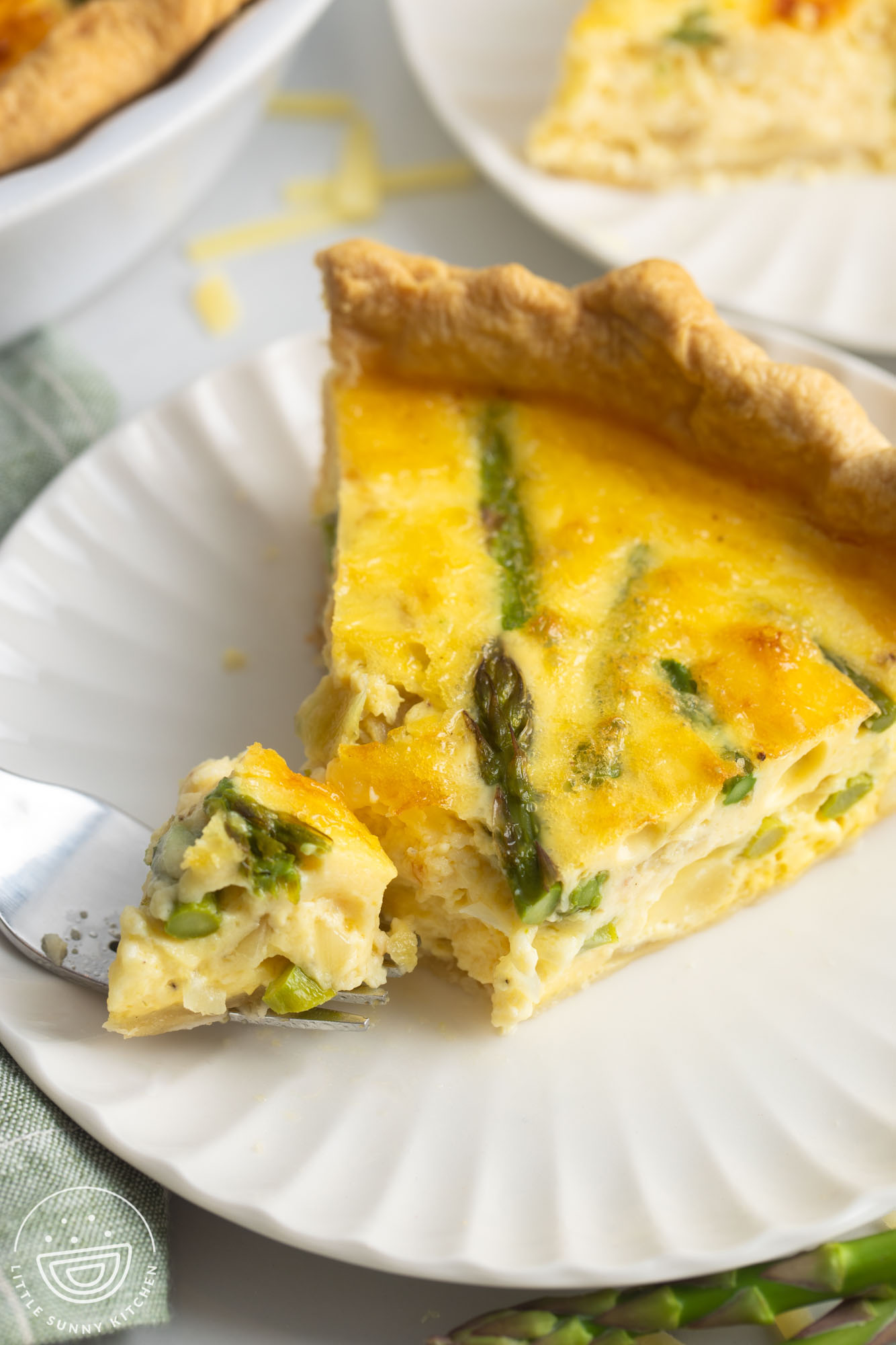 one wedge shaped slice of quiche with asparagus on a small white fluted plate. A fork is taking a bite.