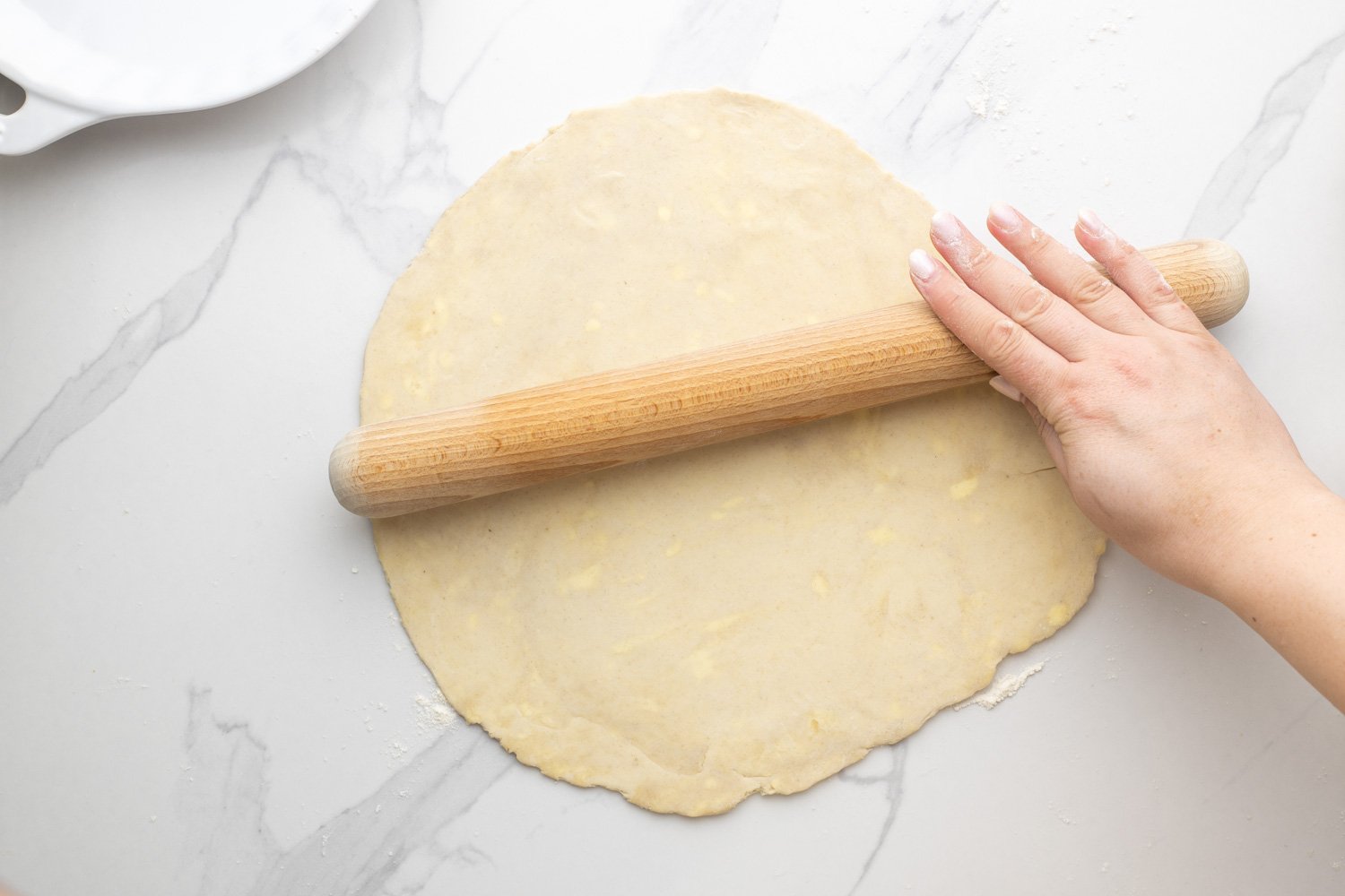 a hand using a straight wooden rolling pin to roll out homemade dough for quiche crust on a marble counter top.