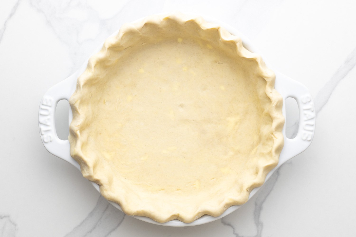 a white ceramic pie pan filled with crust dough that has been crimped around the edges.