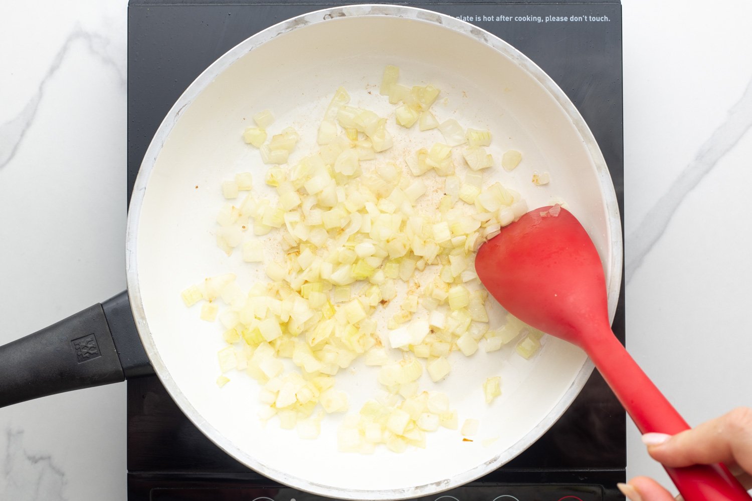 diced onion sauteed in a white skillet over an electric burner, stirred with a red spoon.