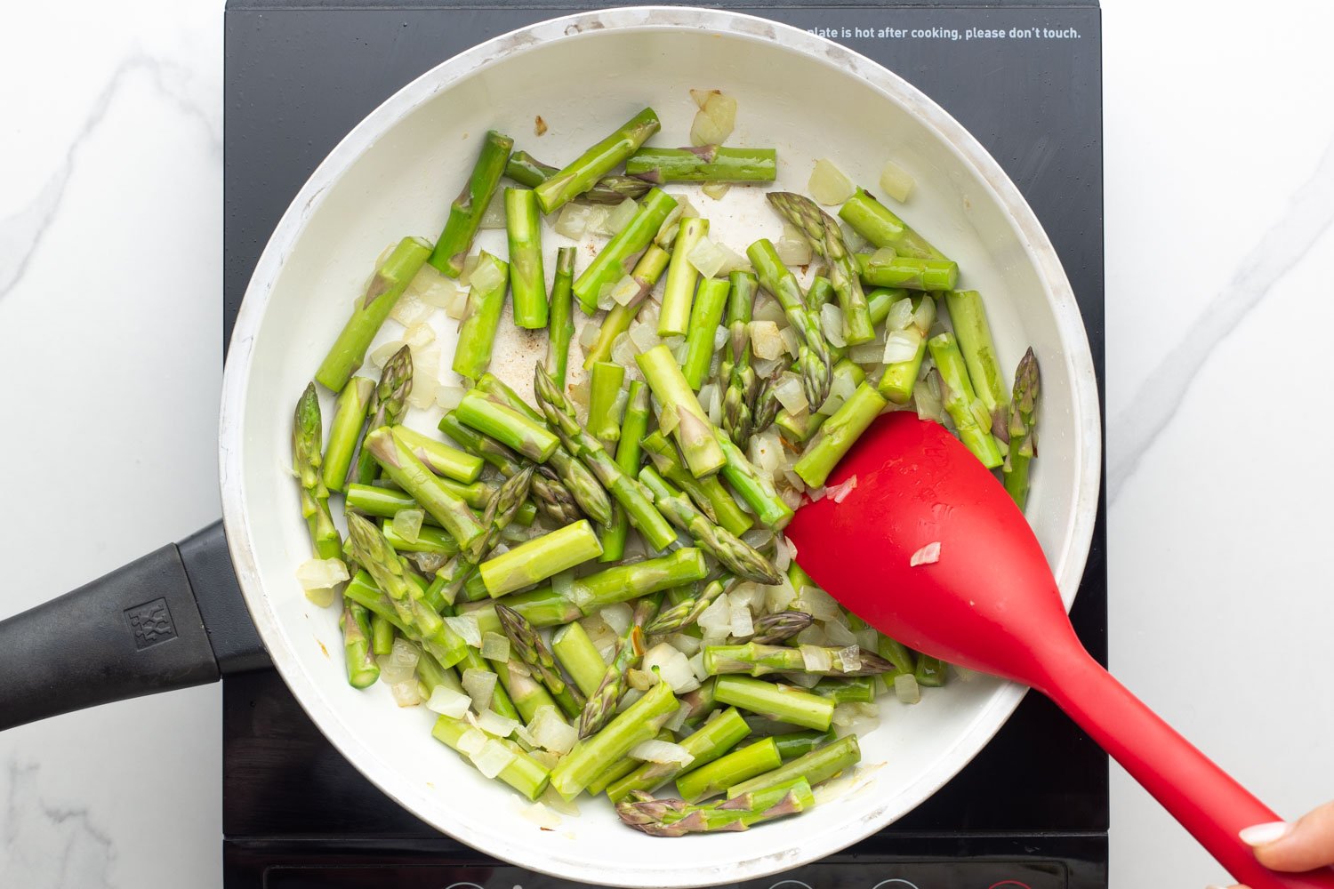 sliced asparagus spears sauteed in a white frying pan with a red spatula.