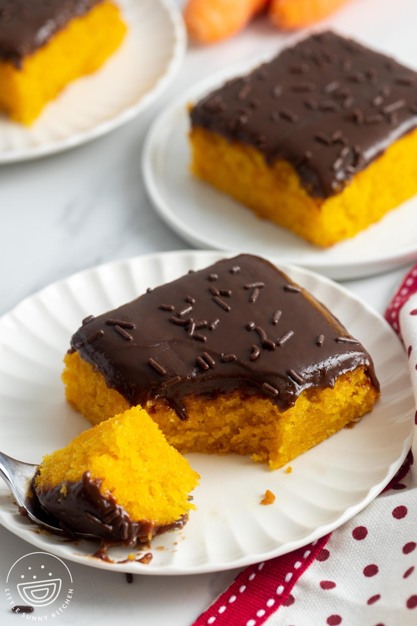 square slices of moist carrot cake with chocolate frosting on small white plates.