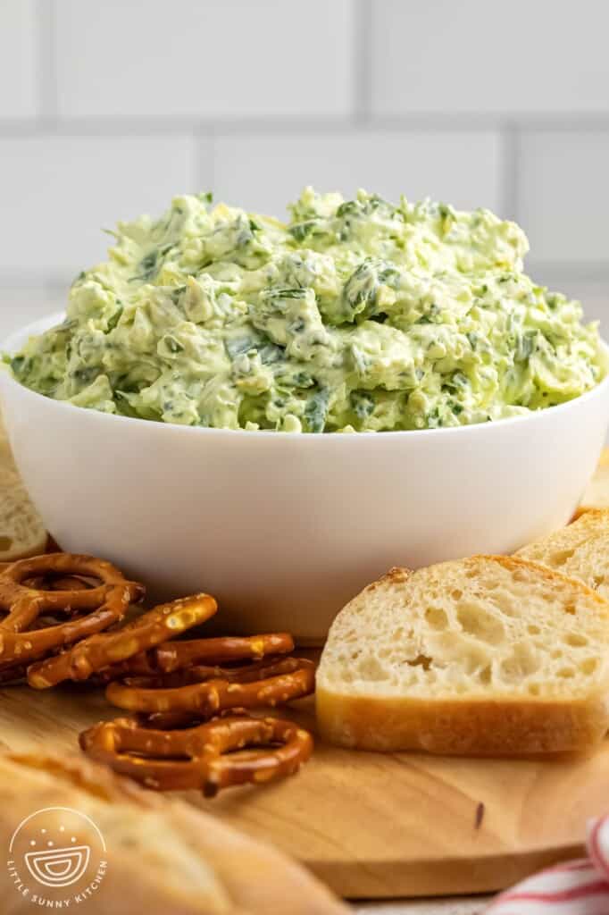 a white bowl of cold spinach and artichoke dip. Next to the bowl are pretzels and small slices of bread.