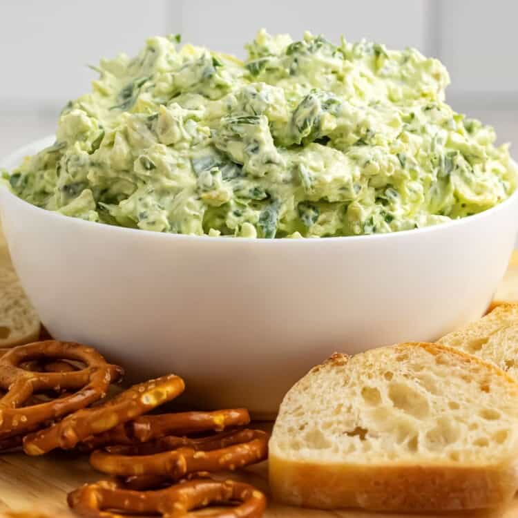 a white bowl of cold spinach and artichoke dip. Next to the bowl are pretzels and small slices of bread.