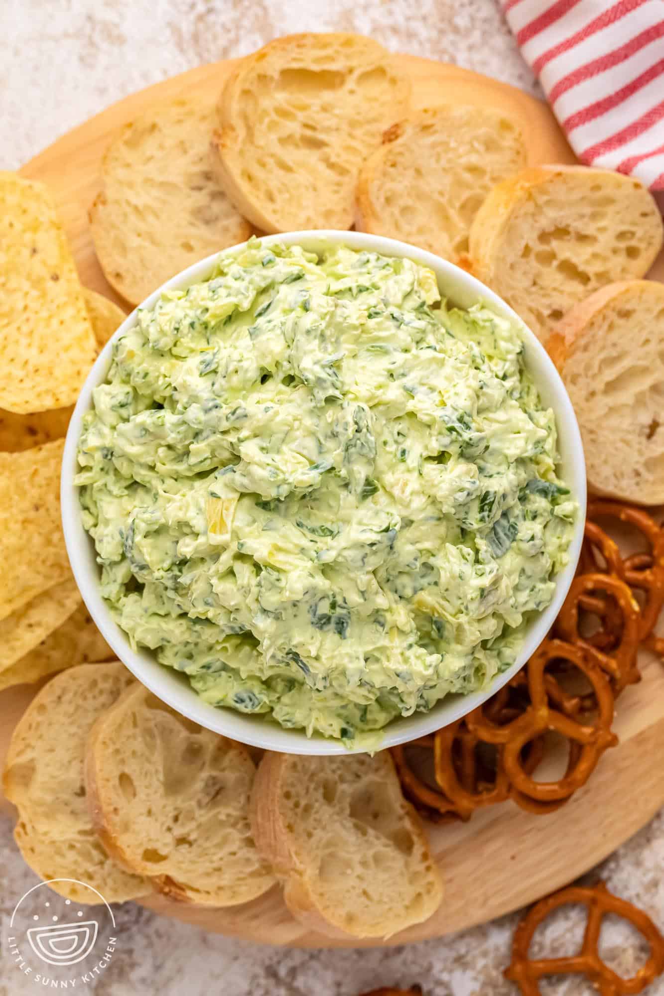 top down view of a bowl of cold spinach artichoke dip surrounded by pretzels, crostini, and tortilla chips