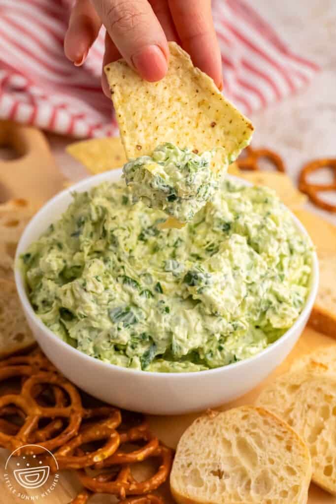 a hand dipping a chip into a bowl of creamy spinach artichoke dip.