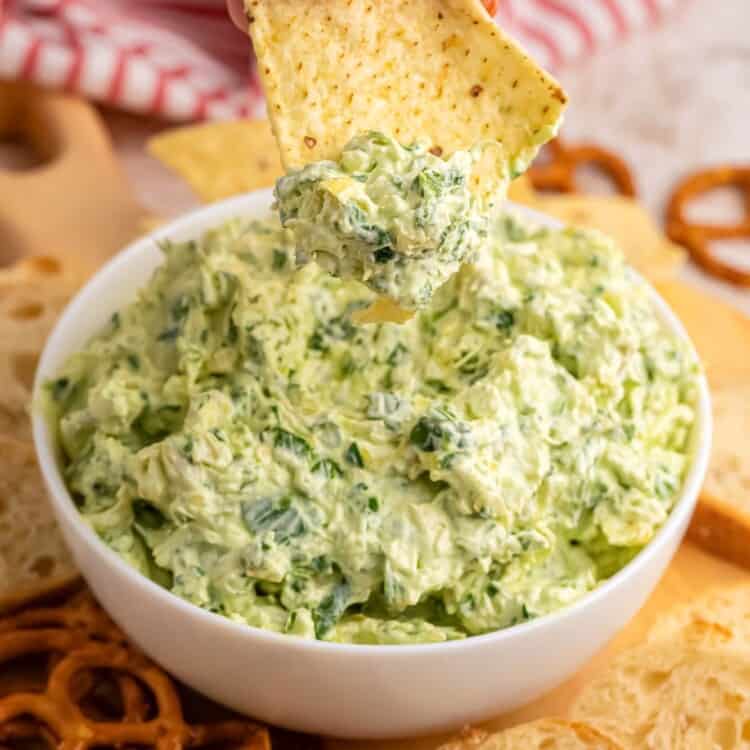 a hand dipping a chip into a bowl of creamy spinach artichoke dip.