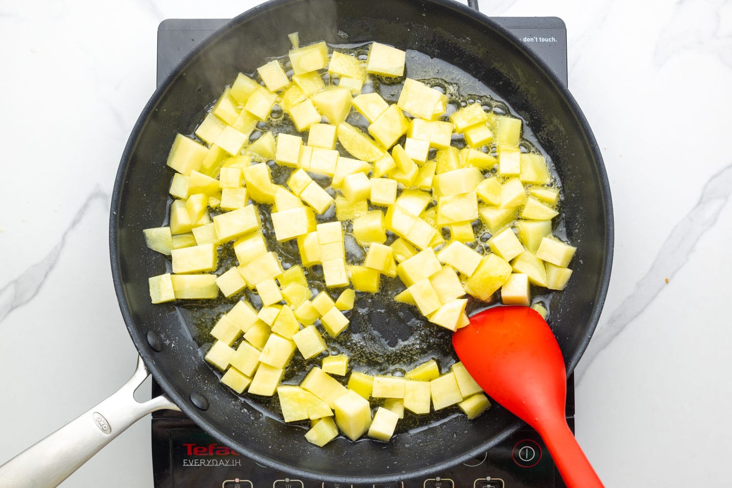 diced potatoes in a non stick skillet with oil, stirred with a red spoon.
