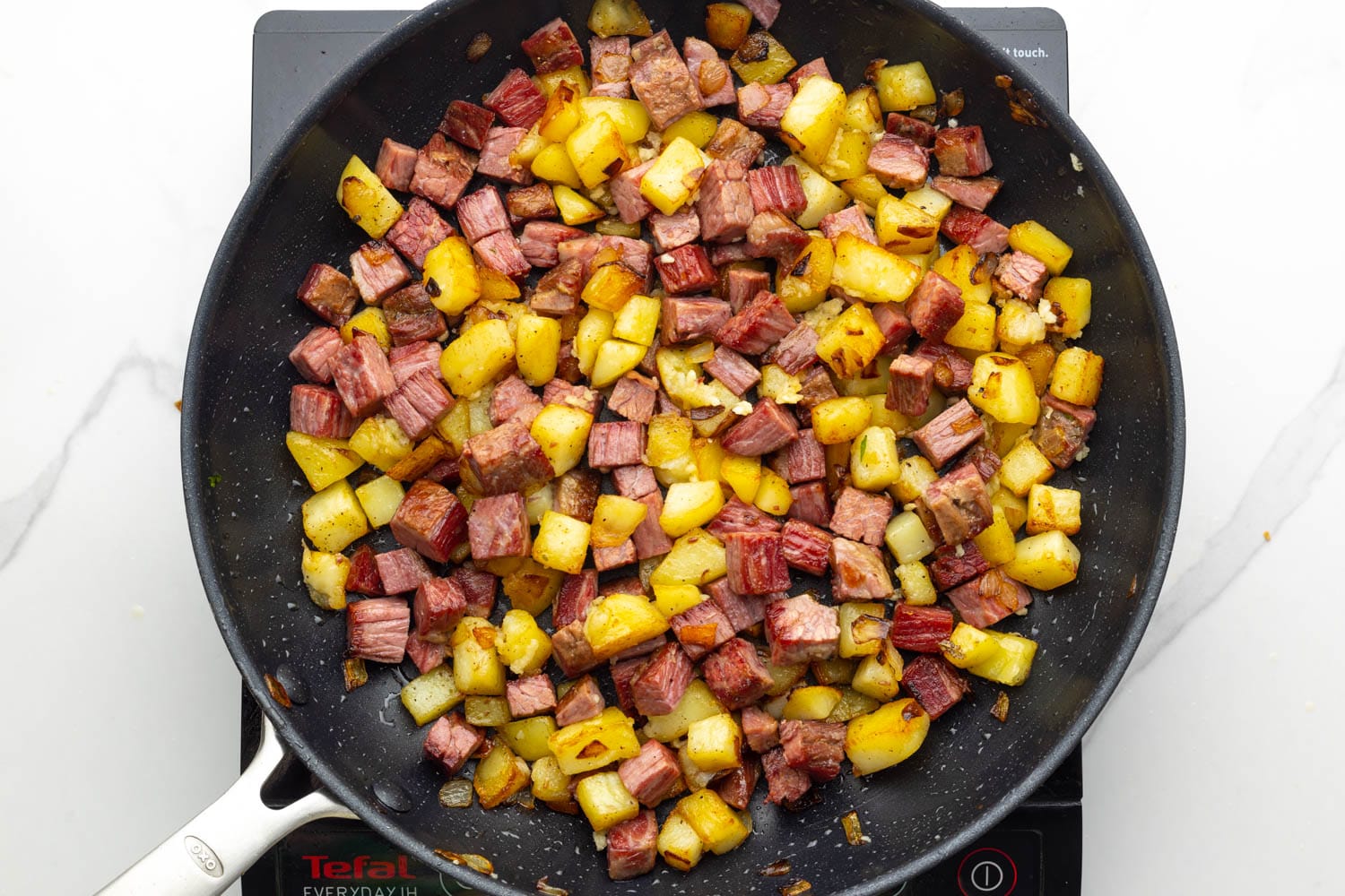 a black nonstick skillet holding browned potatoes an corned beef cubes.