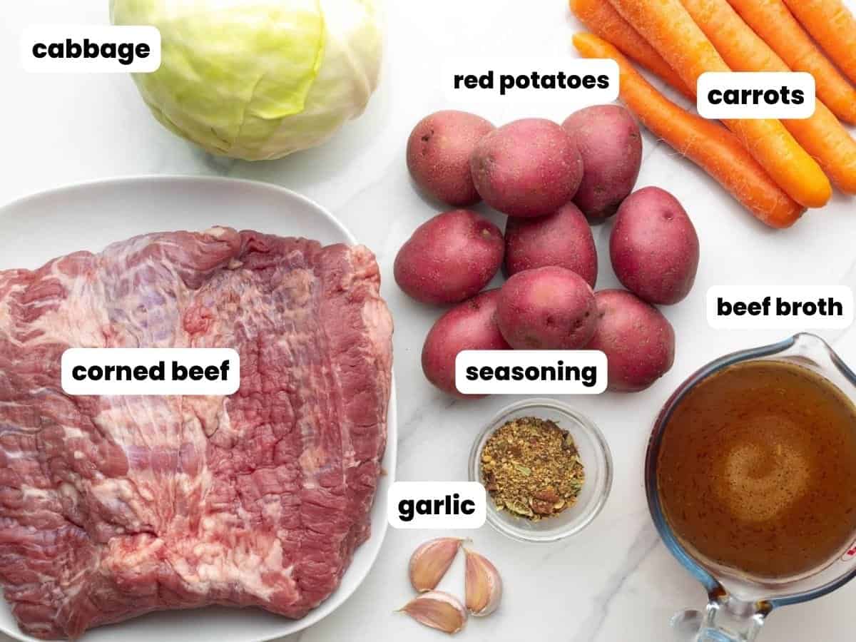 carrots, potatoes, cabbage, a raw corned beef roast, a cut of beef broth, garlic, and seasonings, arranged on a counter.