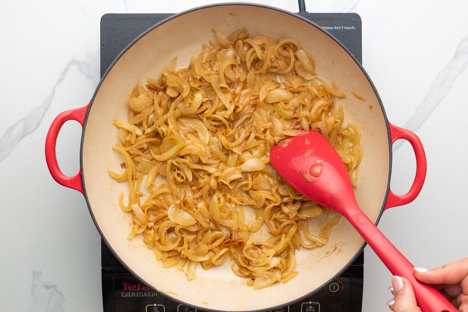 brown caramelized onions in an enameled skillet, stirred with a red spoon.