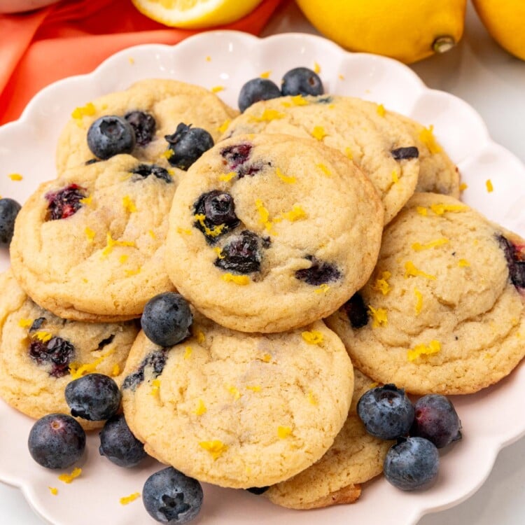 a white fluted plate holding a pile of lemon blueberry cookies, garnished with extra lemon zest and fresh berries.