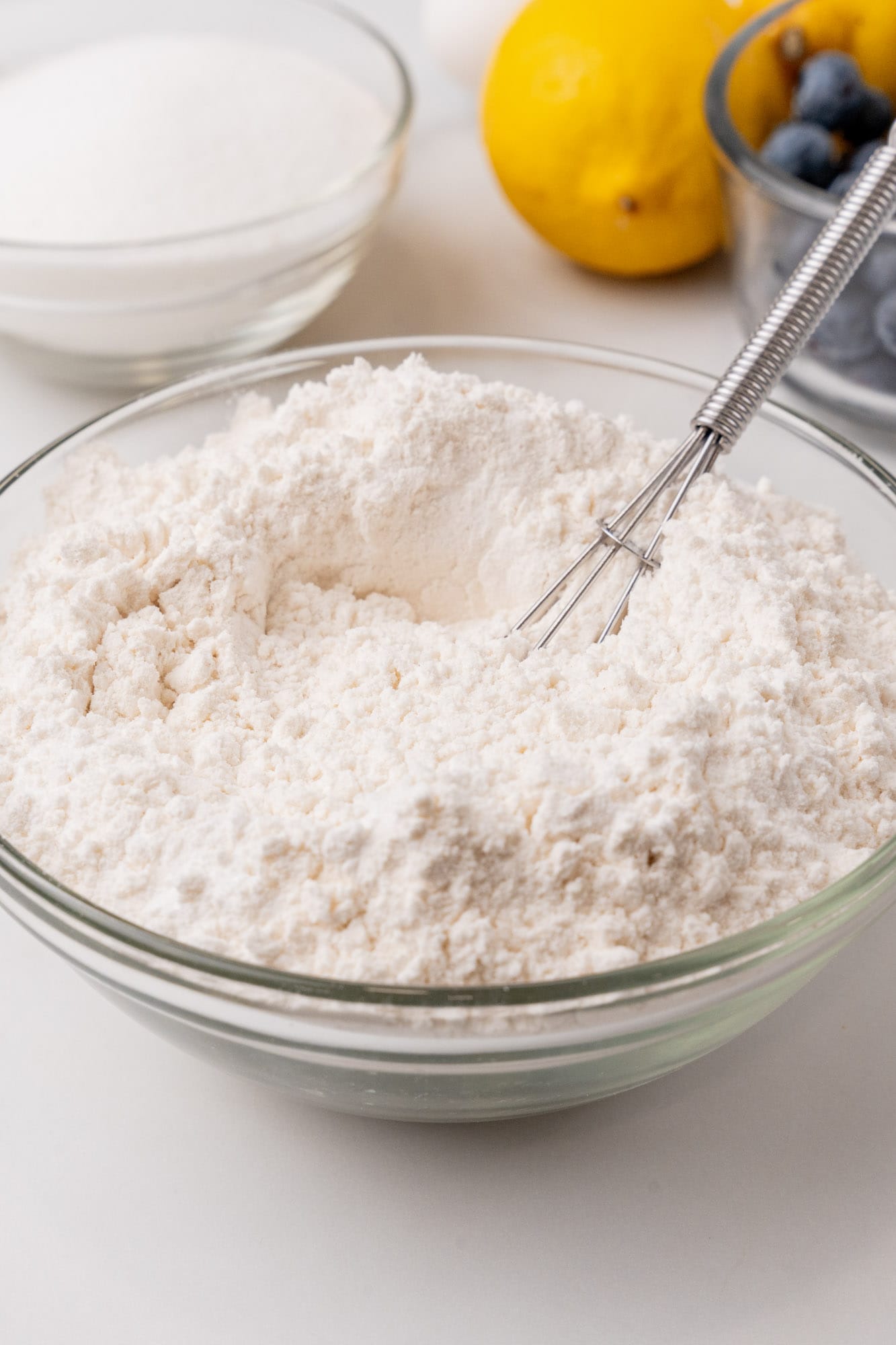 Flour and dry ingredients whisked in a glass bowl. In the background are lemons, sugar, and blueberries.