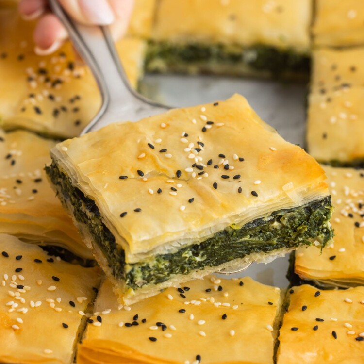 a hand holding a square piece of spanakopita with a silver spatula.