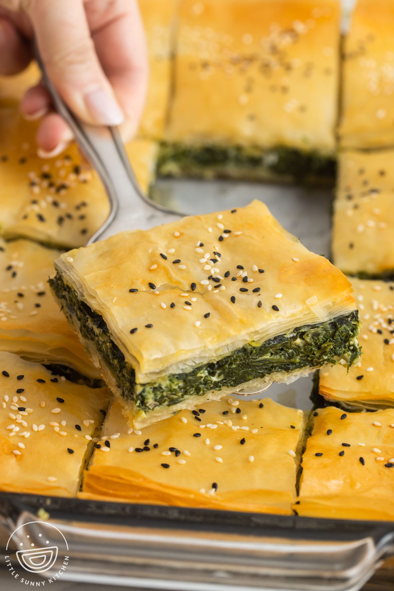 a hand holding a square piece of spanakopita with a silver spatula.