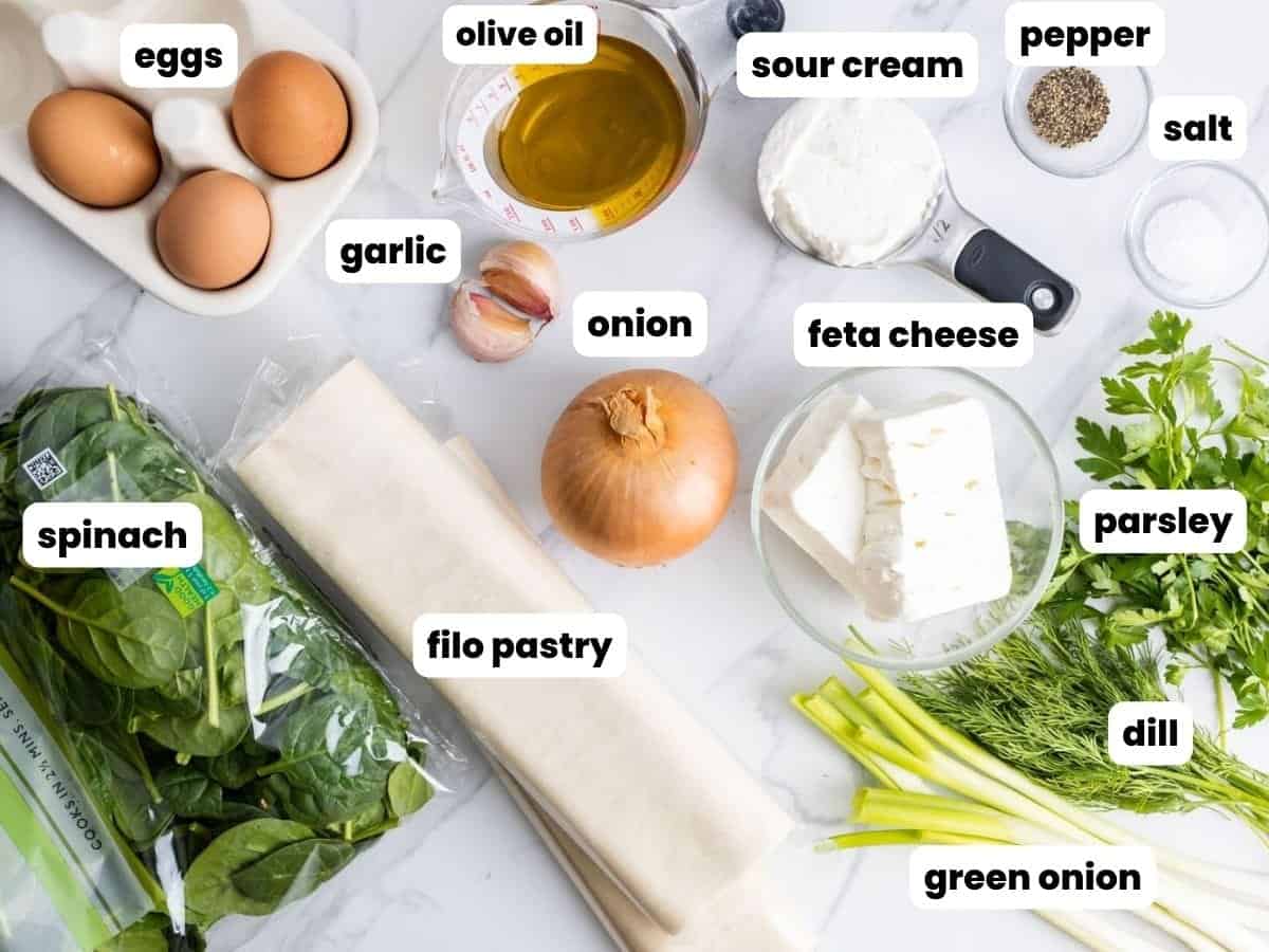 The ingredients needed to make homemade spanakopita including filo pastry, feta, and spinach, arranged on a marble counter.