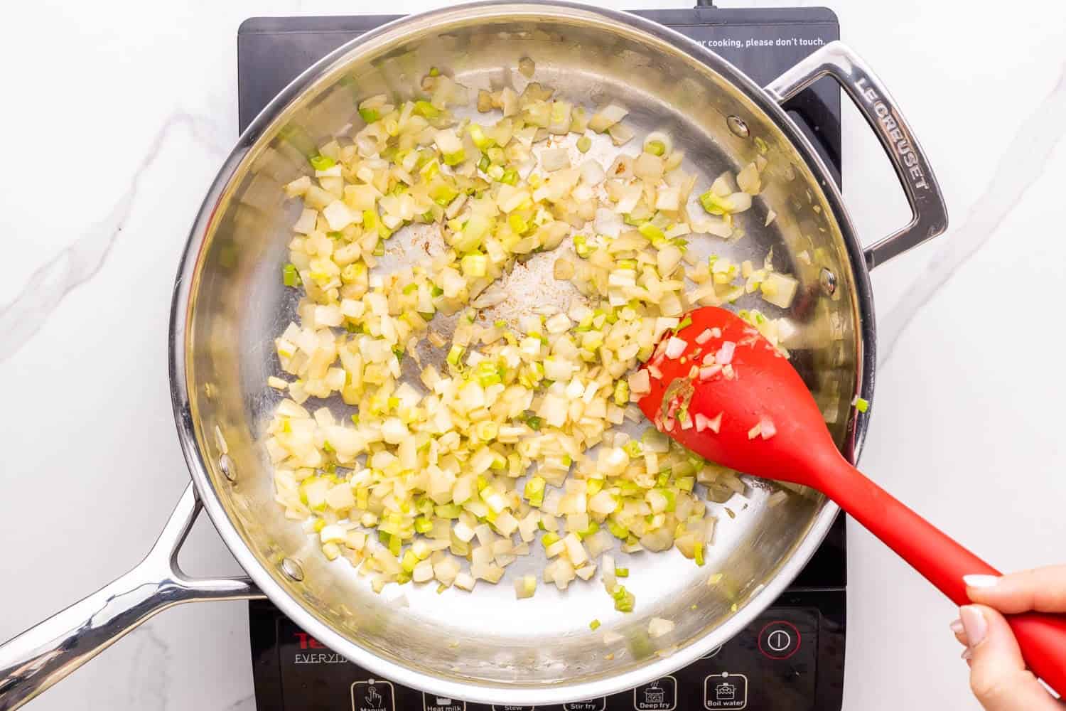 a stainless steel skillet on an electric burner. In the pan are sauteed onions, stirred with a red spoon.