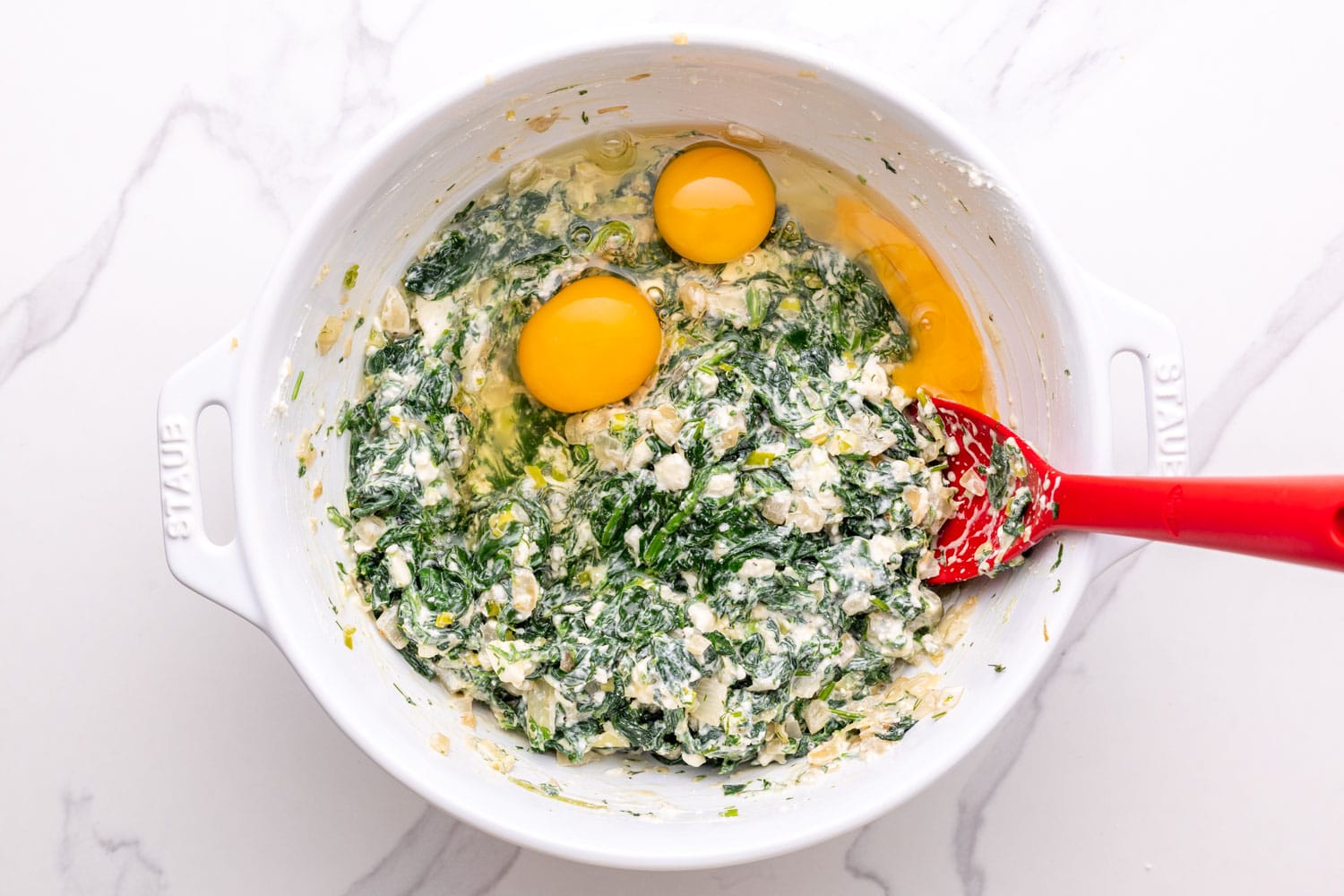 eggs and cheese added to a white bowl of sauteed spinach to make greek spinach pie