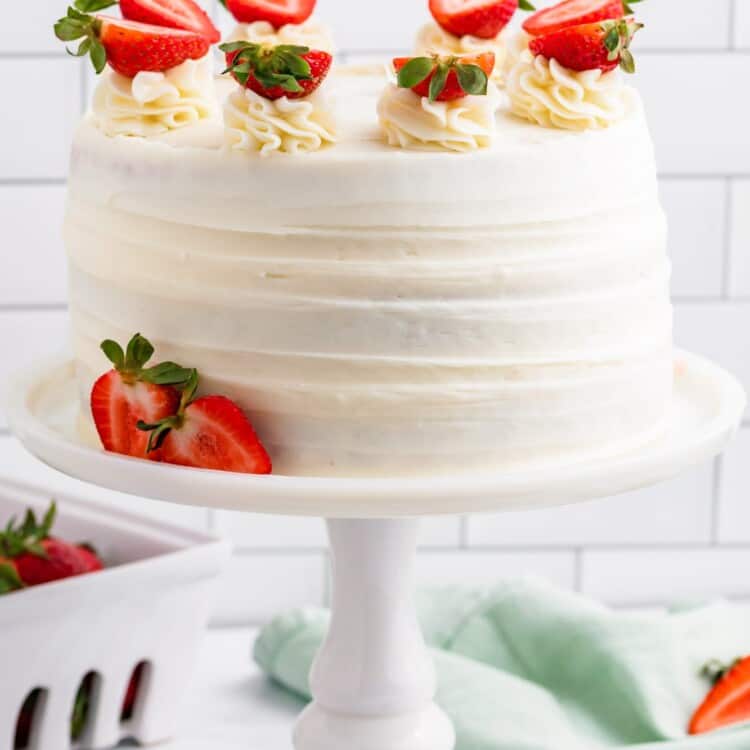 a white ceramic cake pedestal holding a round strawberry cake frosted with cream cheese frosting and topped with fresh strawberries that have been cut in half.