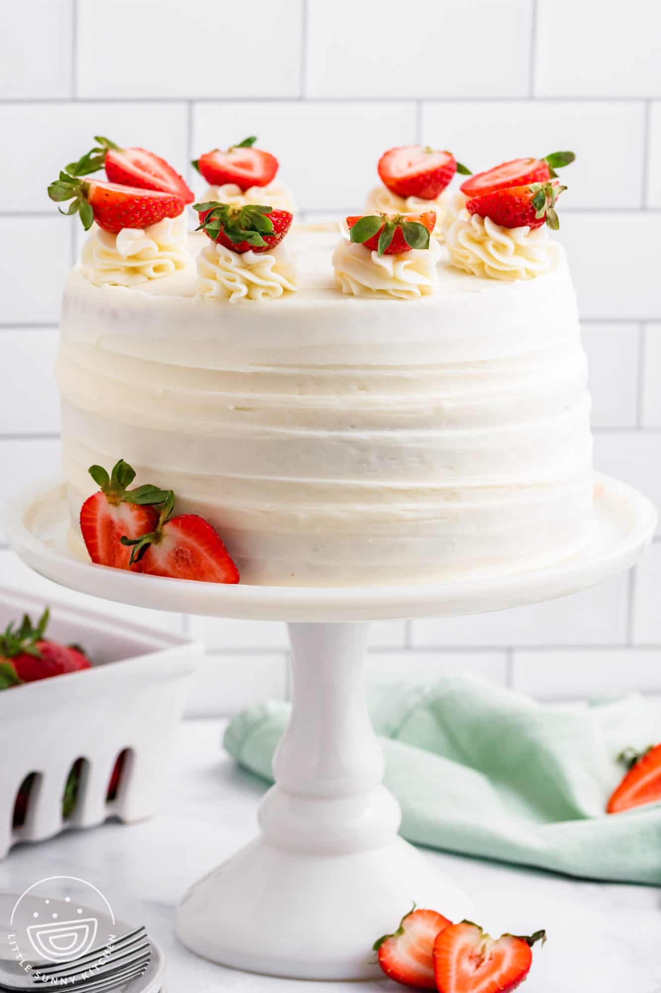 a white ceramic cake pedestal holding a round strawberry cake frosted with cream cheese frosting and topped with fresh strawberries that have been cut in half.