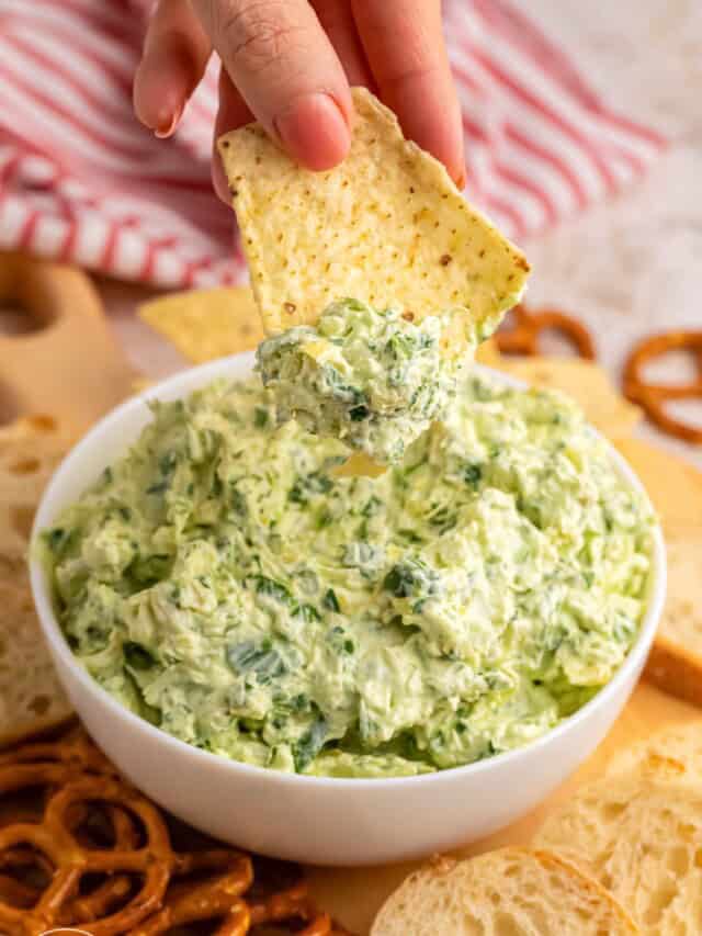 a hand dipping a chip into a bowl of creamy spinach artichoke dip.