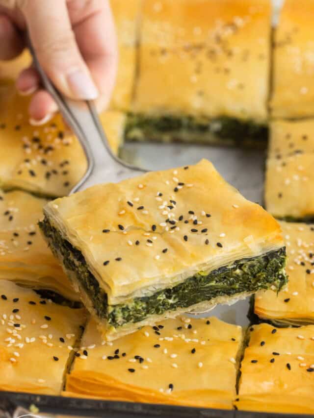 a hand holding a square piece of spanakopita with a silver spatula.