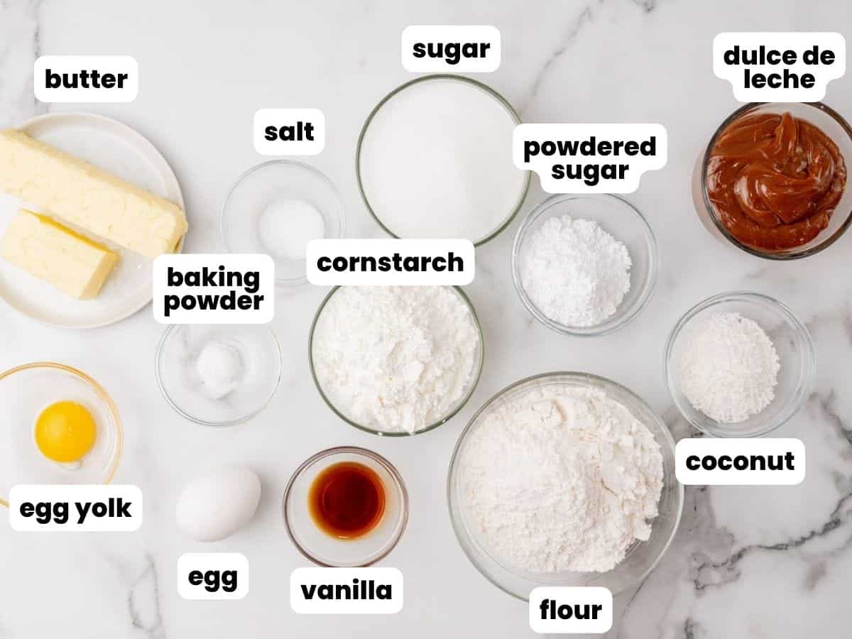 the ingredients for alfajores, including dulce de leche, all measured into small bowls and arranged on a marble counter.