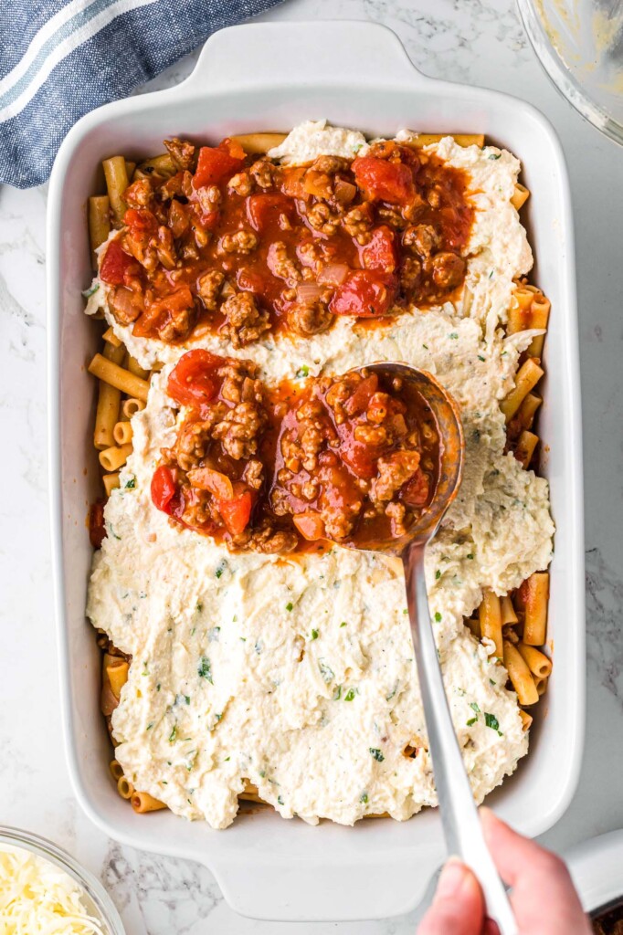 a large spoon adding meat sauce to a casserole dish of pasta and ricotta cheese.