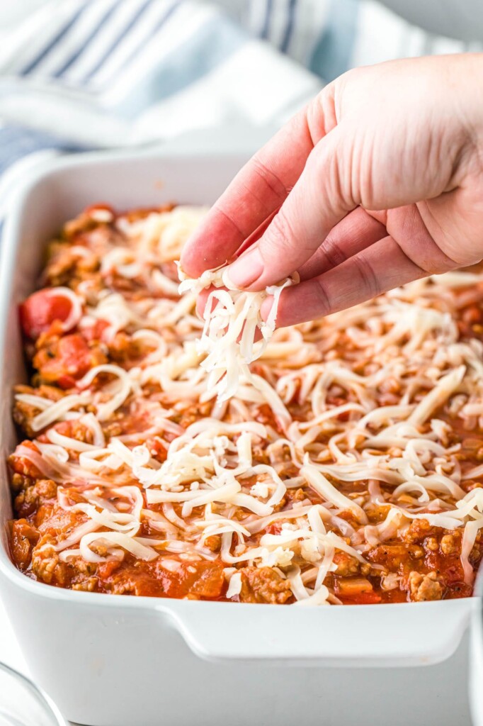 a hand sprinkling grated mozzarella cheese over baked ziti with sausage and tomatoes