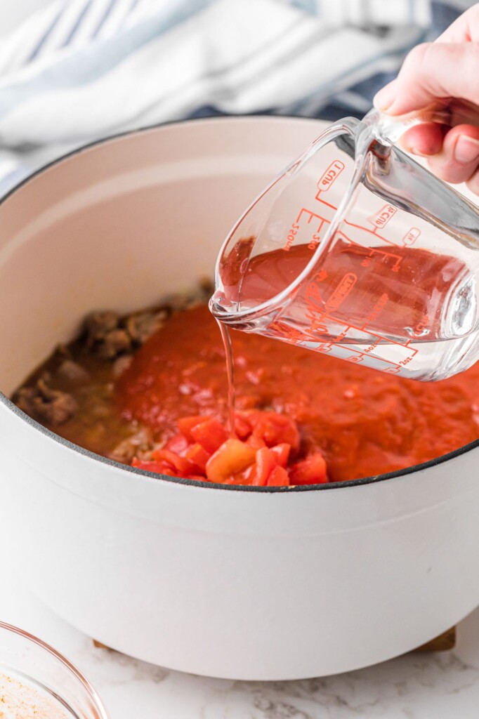 a hand pouring water into tomato sauce in a dutch oven using a glass measuring cup.