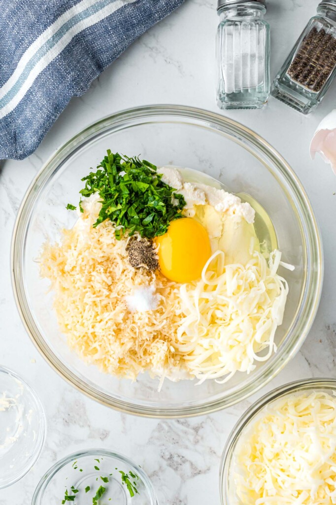 a glass mixing bowl holding ricotta cheese, chopped herbs, one raw egg, shredded mozzarella and grated parmesan.