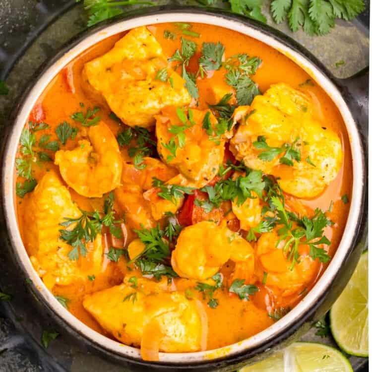 a large bowl of creamy orange colored fish stew on a black platter of lime wedges, garnished with cilantro.