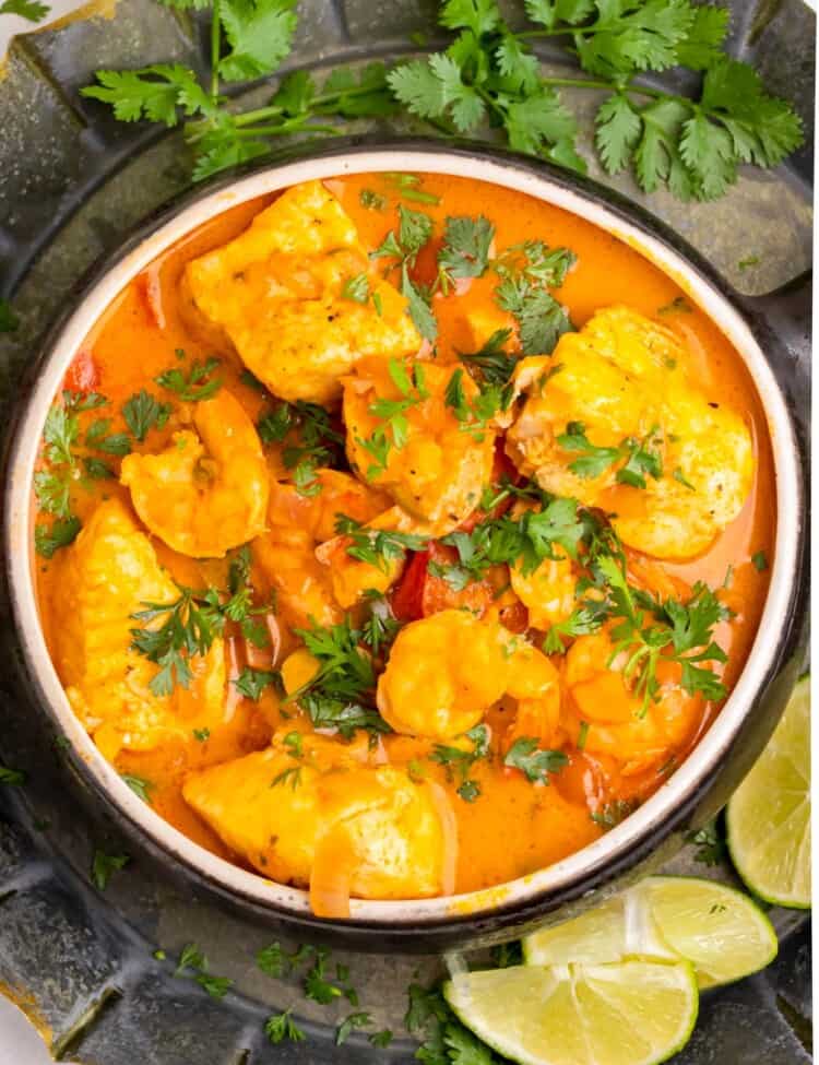 red stew with fish and shrimp, garnished with cilantro. The bowl is on a black platter with herbs and lime wedges.