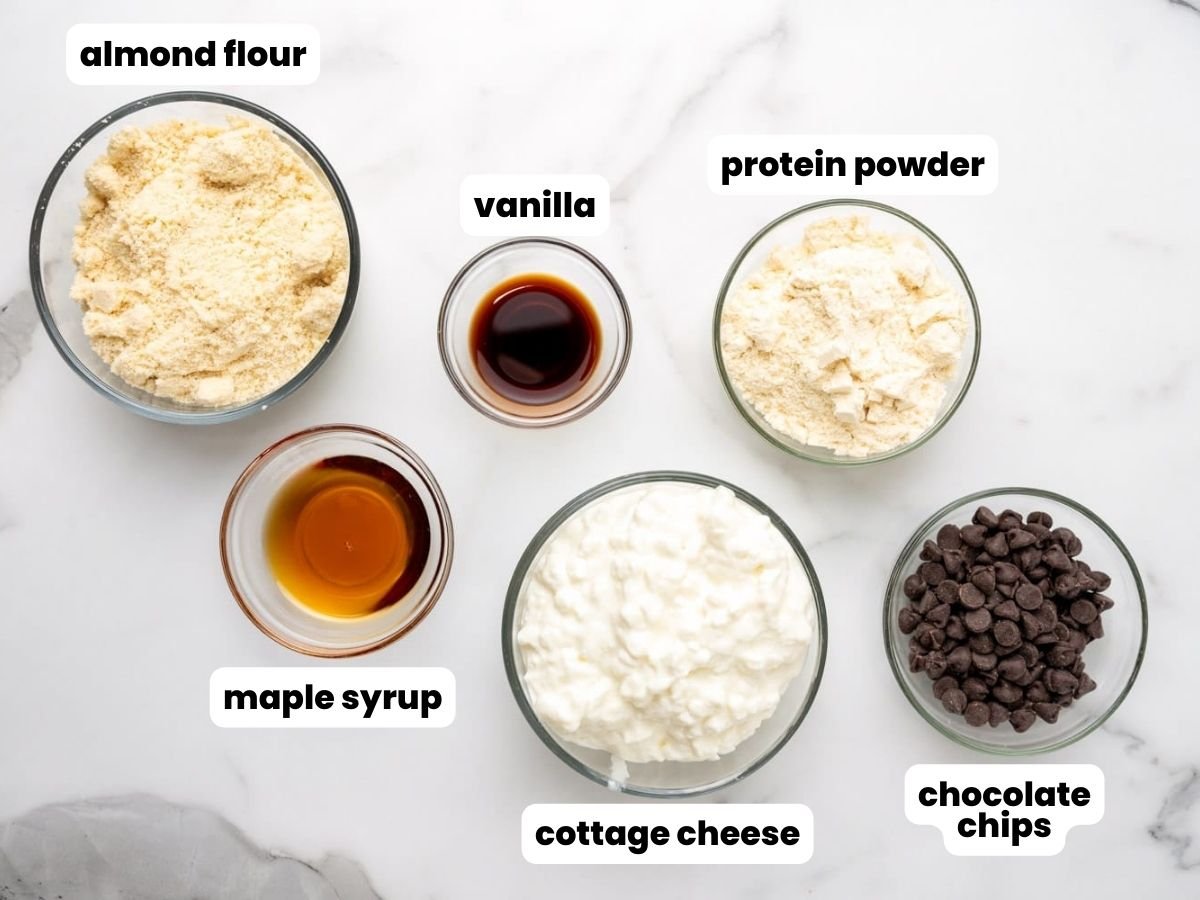 The ingredients needed to make high protein cookie dough with cottage cheese and protein powder, all measured into small glass bowls and arranged on a marble counter.