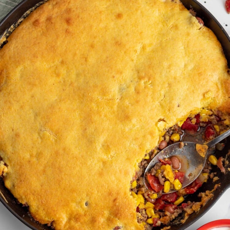 cowboy cornbread casserole in a large cast iron skillet. A spoon has removed one serving from the side of the pan.