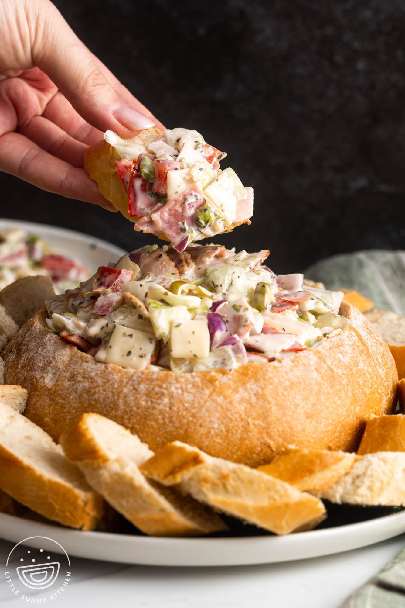 a hand holding a sliced baguette with italian hoagie dip over a large bread bowl.