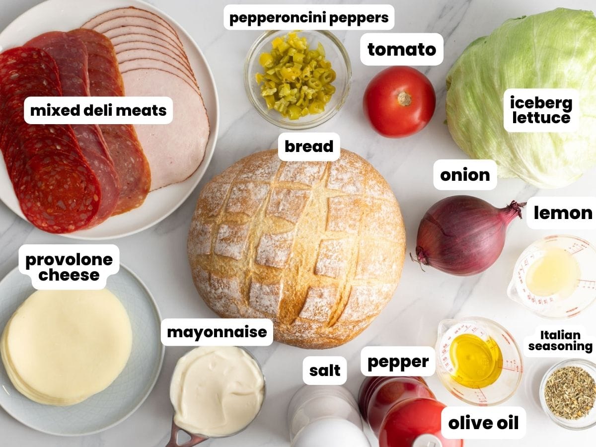 a platter of mixed deli meats next to a round loaf of bread, provalone cheese slices, lettuce, tomato, onions, and other ingredients needed to make Italian Hoagie Dip