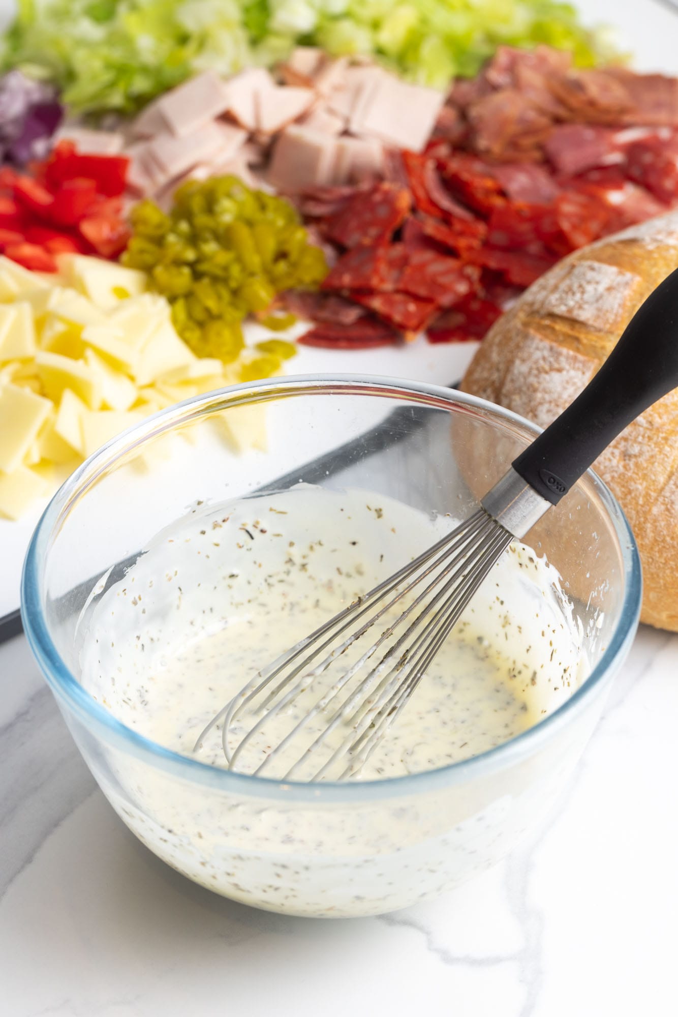 creamy dressing for Italian hoagie dip in a small bowl with a whisk next to a cutting board of chopped meats, cheeses, and vegetables.