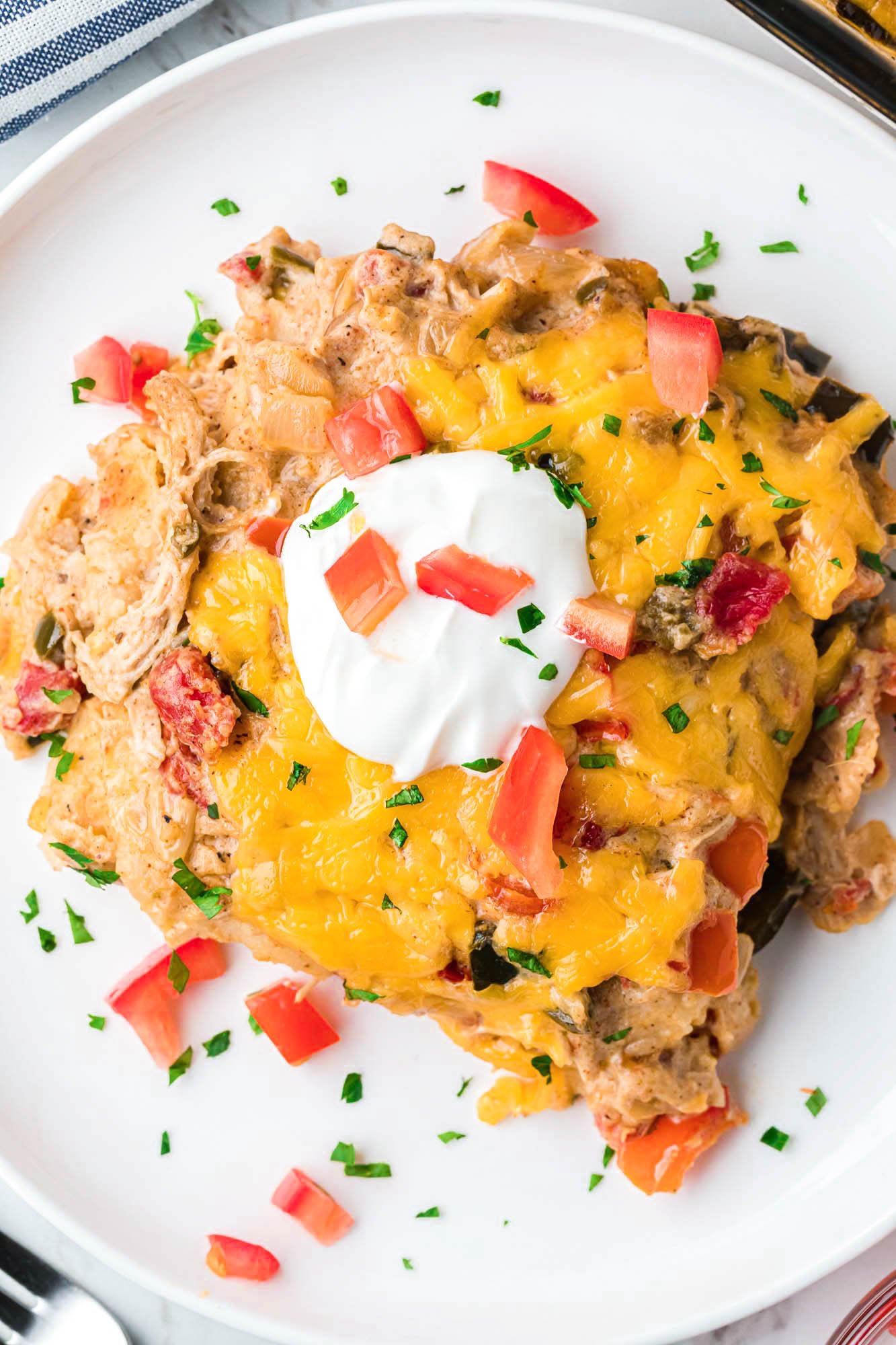 a square serving of king ranch casserole on a plate, topped with sour cream and diced tomatoes. 