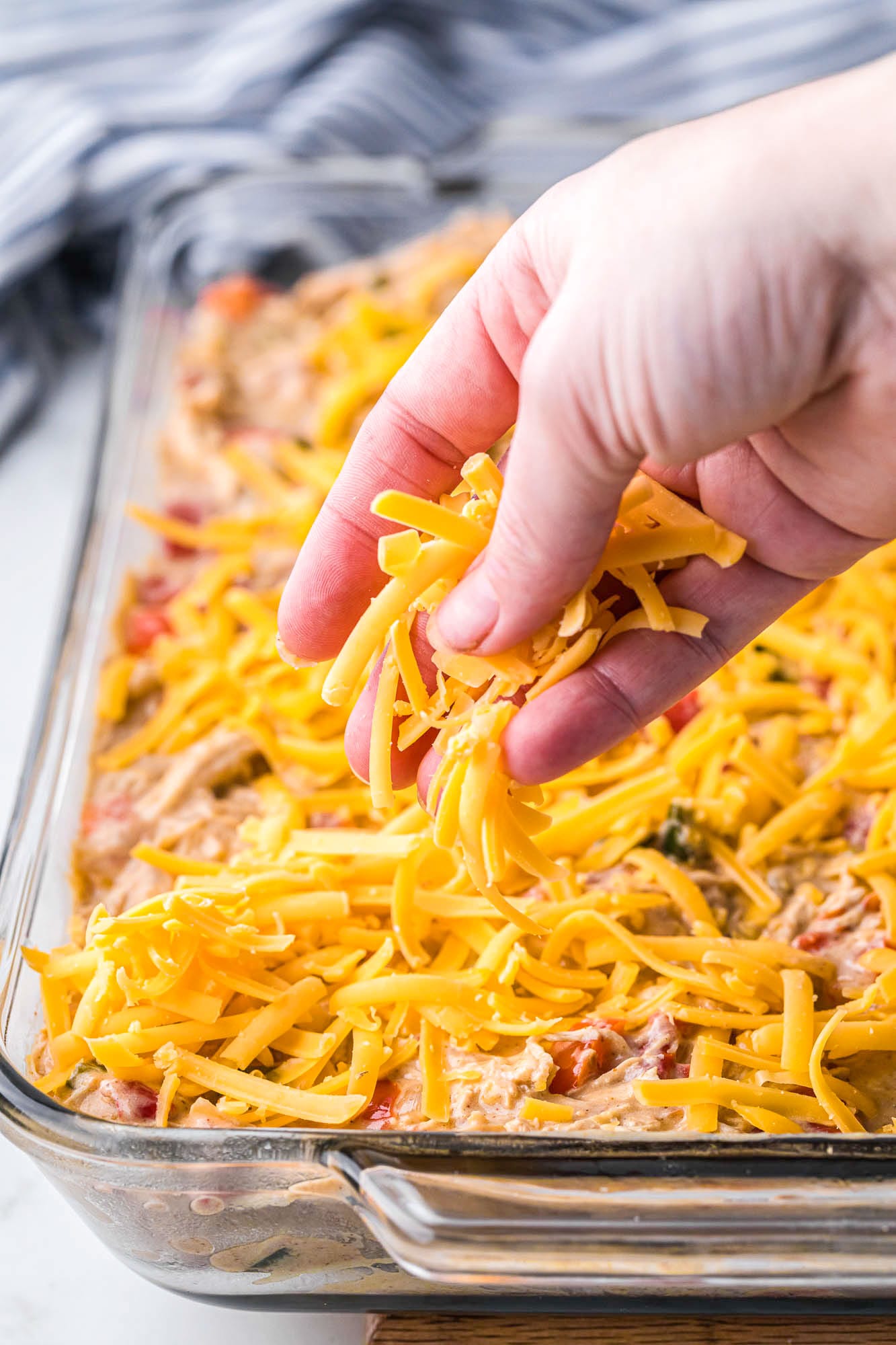 a hand adding a final layer of shredded cheddar cheese to a casserole. 