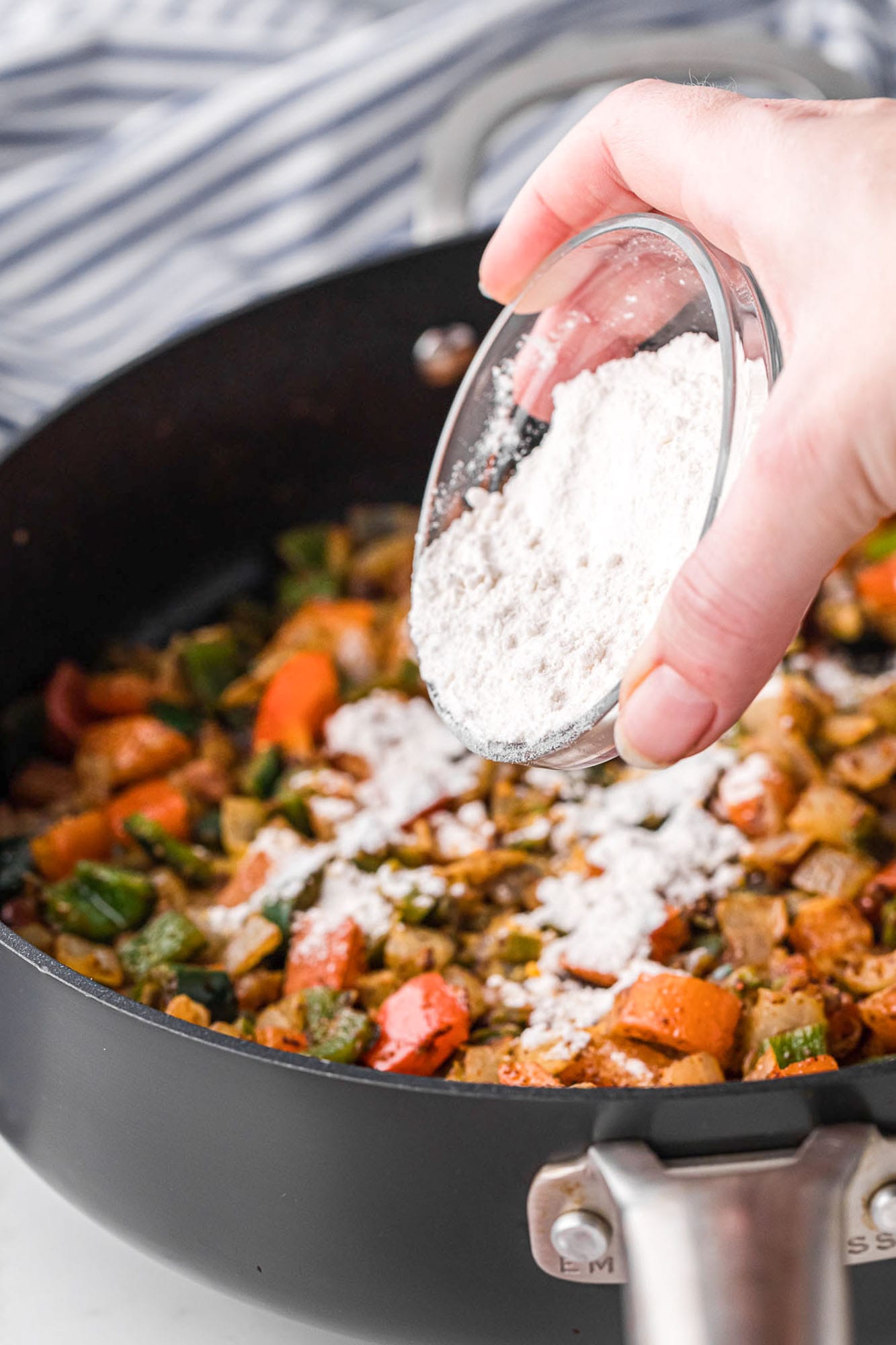 a hand holding a small bowl of flour, sprinkling it over cooked and seasoned peppers and onion in a large skillet. 
