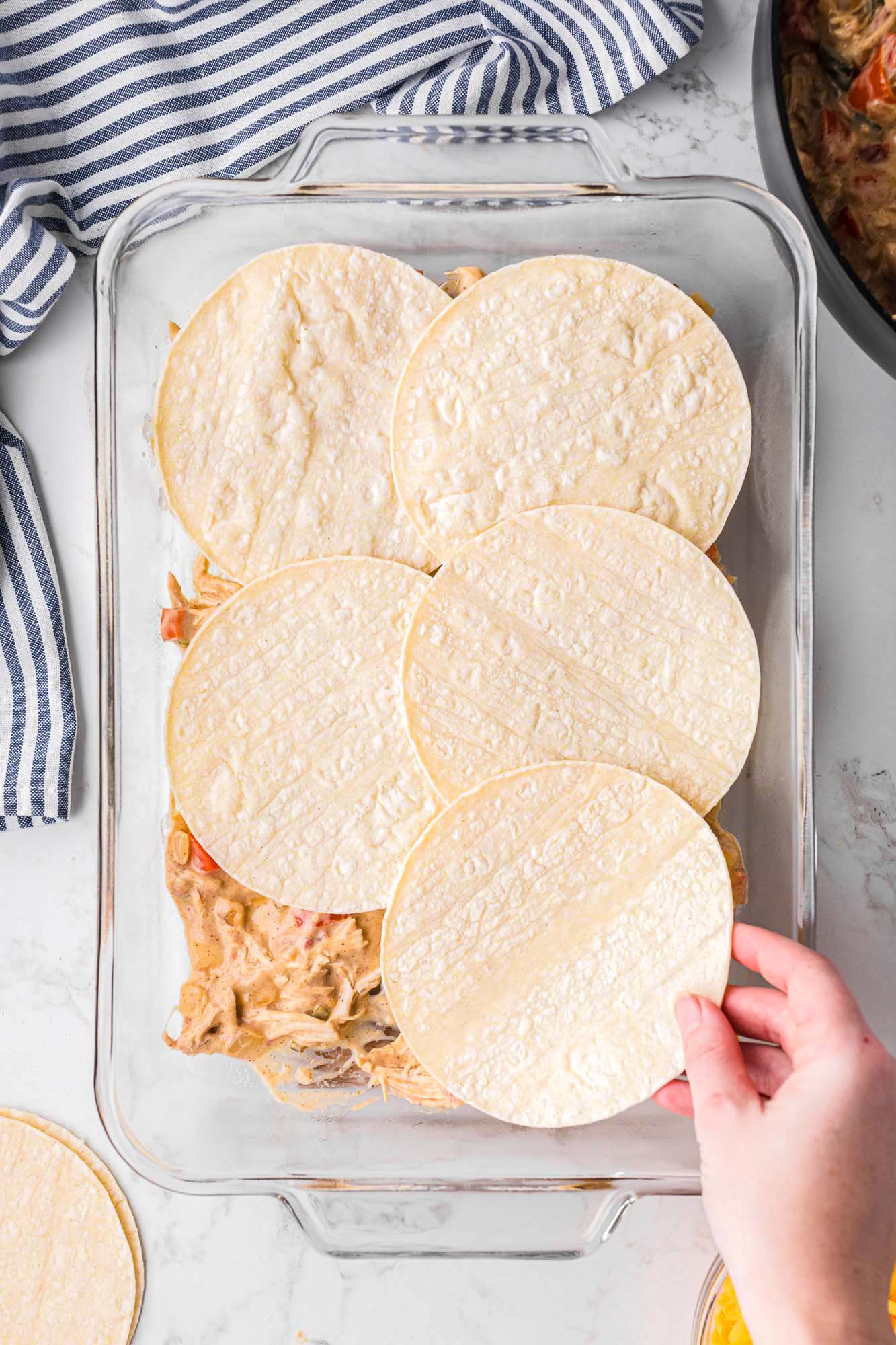 flour tortillas layered over shredded chicken filling in a glass casserole dish. 